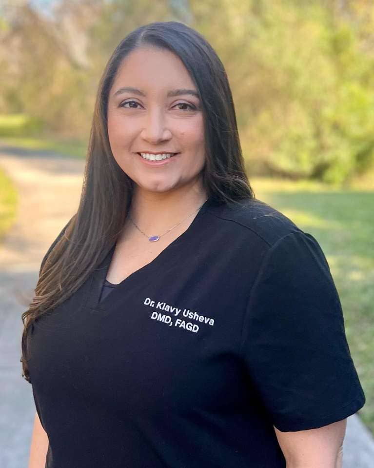 Klavy Usheva, DMD | Dr. Klavy Usheva, DMD, FAGD, smiling outdoors in a black scrub top embroidered with her name.
